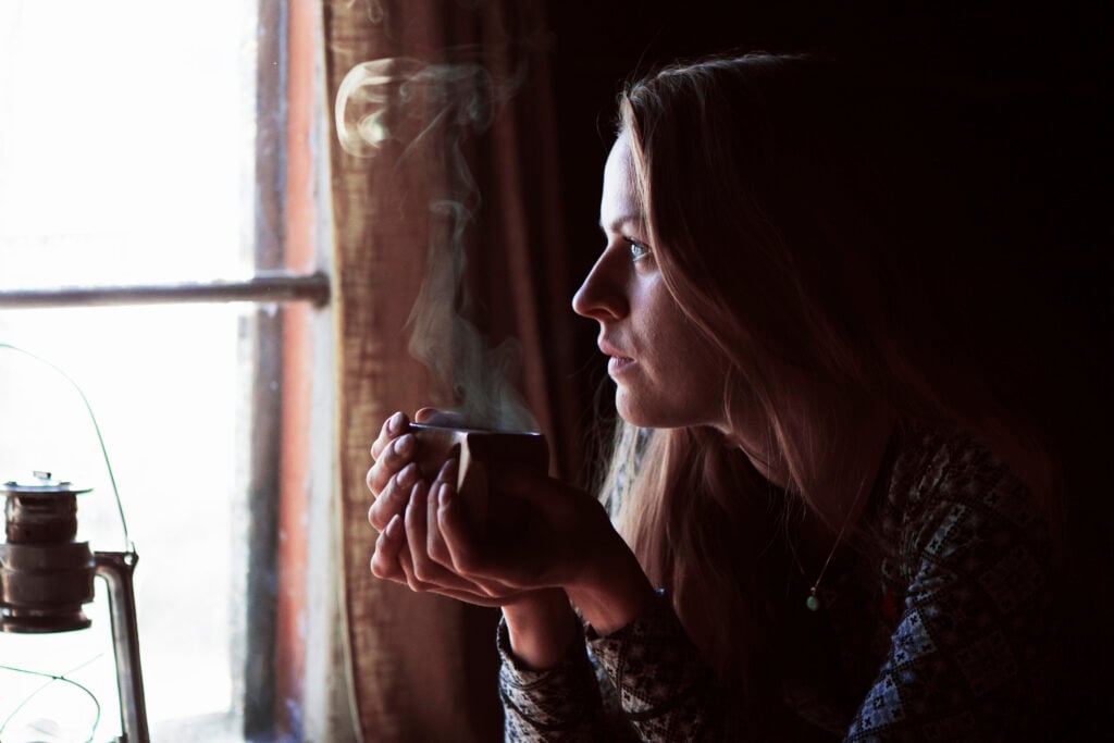 Nainen istuu hämärässä huoneessa ja katsoo ulos ikkunasta höyryävä kuppi käsissään. Pehmeä valo lankeaa hänen kasvoilleen, ja vieressä näkyy vanha öljylamppu.